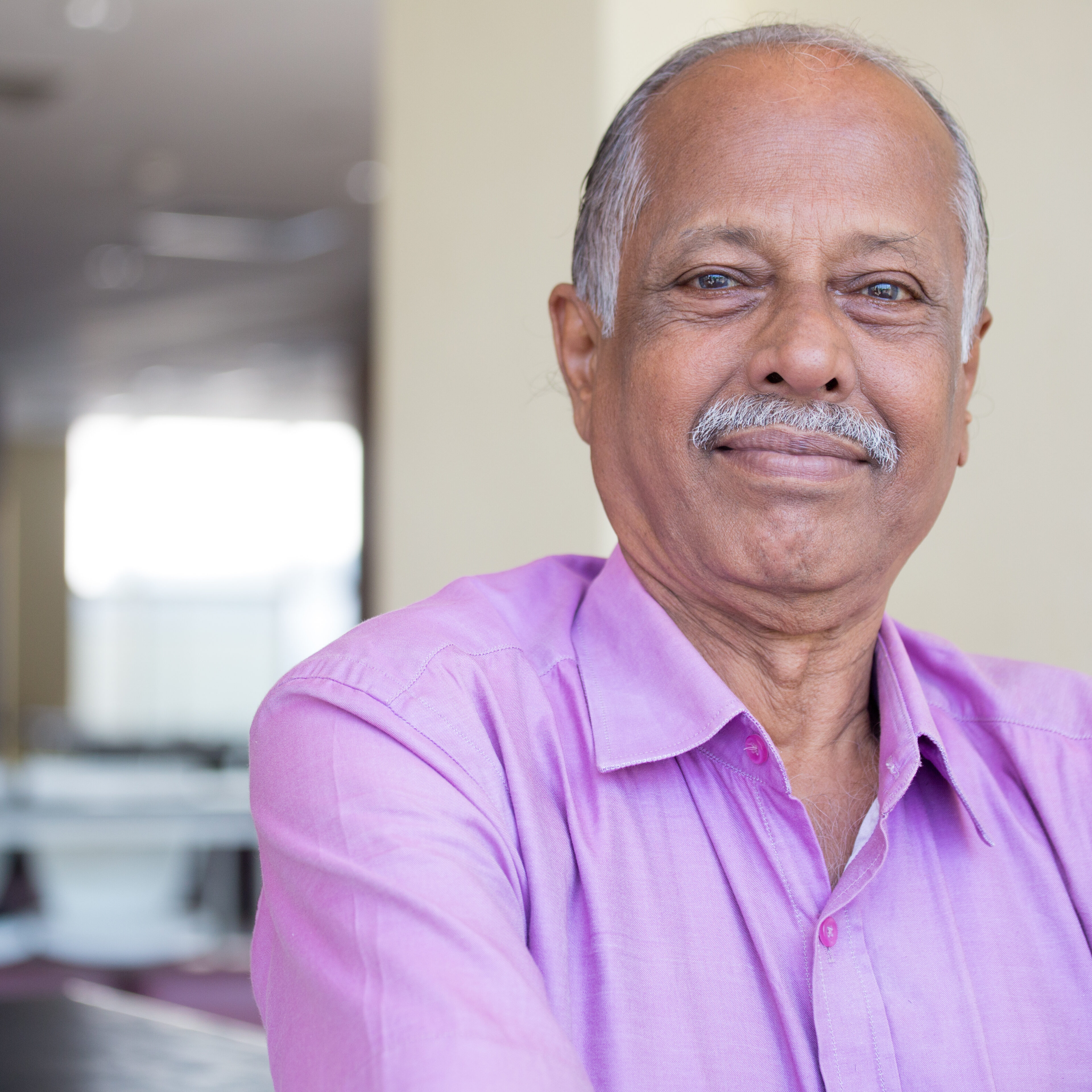 Closeup headshot portrait of elderly gentleman sitting down in pink shirt smiling, content, isolated indoors white wall background